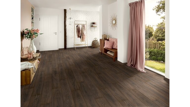 Heller Raum mit dunklem Holzboden, weißen Wänden, Möbeln und Gardine, die auf einen Garten blicken.